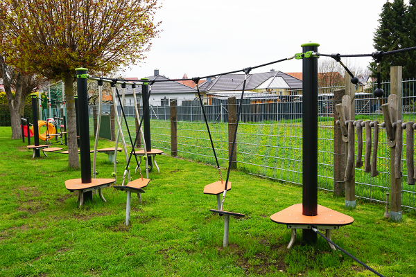 Balancierparcours mit verschiedenen schwingenden Trittplattformen auf einem grünen Kita-Außengelände, im Hintergrund ein Kletterelement aus Holz.