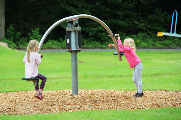 Zwei Mädchen spielen an einem rotierenden Hip-Hop-Spielgerät mit zwei gegenüberliegenden Sitzen auf einer Wiese mit Fallschutzbelag.