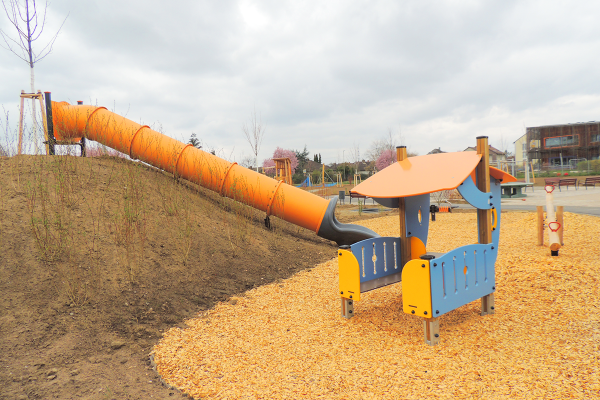 Eine lange orange Tunnelrutsche führt von einem Hügel hinab auf eine Fallschutzfläche mit einem bunten Spielhaus im Generationenpark Philippsburg.