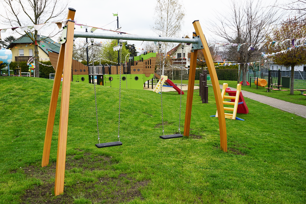 Doppelschaukel aus Holz mit zwei schwarzen Sitzen auf einer Wiese, im Hintergrund ein großes Spielschiff und weitere Spielgeräte auf dem Kita-Spielplatz.