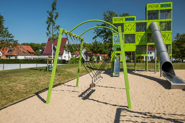 Grüne Tampenschaukel mit dickem Seilbalken auf Sandfläche, im Hintergrund ein großer Kletterturm mit Edelstahlrutsche und Rutschelement im Stil eines Förderturms.