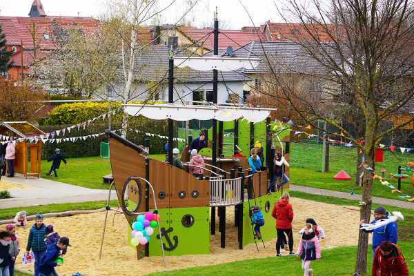 Mehrere Kinder spielen aktiv auf und um ein großes Spielschiff auf einem Kita-Spielplatz, umgeben von bunten Wimpeln und Luftballons.