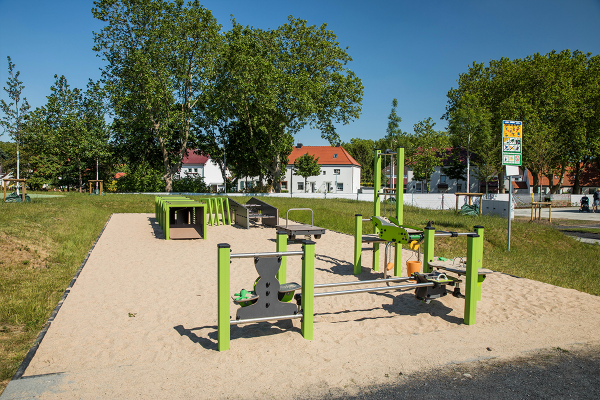 Sandspielbereich mit grünen Spielstationen zum Schütten, Drehen und Fördern von Sand sowie einem Kriechtunnel. Die Fläche ist mit hellem Spielsand bedeckt.