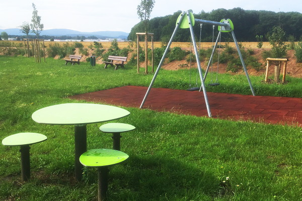 Aufenthaltsbereich mit Jugendbank, Tisch und Hockern sowie Doppelschaukel auf dem Spielplatz Eisenach-Hötzelsroda in naturnaher Umgebung.