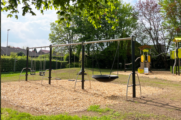 Schaukelkombination mit Nestschaukel, Kleinkindersitzen und klassischen Schaukelsitzen auf Holzschnitzel-Fallschutz in grüner Parkumgebung.