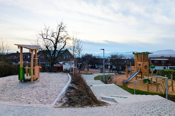 Übersicht eines Naturholz-Spielplatzes in Hanglage mit Sandbereich, Spielturm mit Rutsche und barrierearmen Wegeflächen in Eislingen.