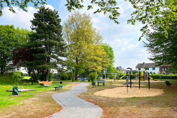 Neugestalteter Quartiersspielplatz mit geschwungenem Weg, Spielbereichen, Sitzgelegenheiten und naturnaher Einbettung in eine grüne Parklandschaft.