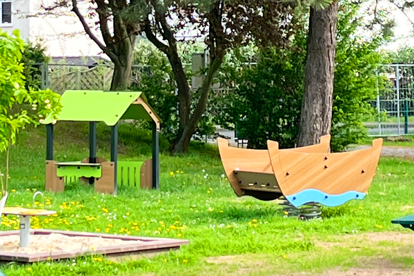 Kleinkindbereich mit Spielhäuschen, Federspielgerät in Bootsform und Sandspielfläche, eingebettet in eine grüne, baumbestandene Umgebung.
