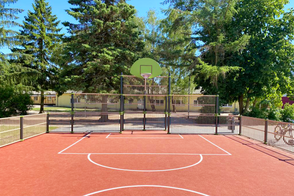 Basketballspielfeld einer Multisportanlage auf dem Schulhof mit rotem Belag, Korb und umlaufender Metallumzäunung vor Baumbestand