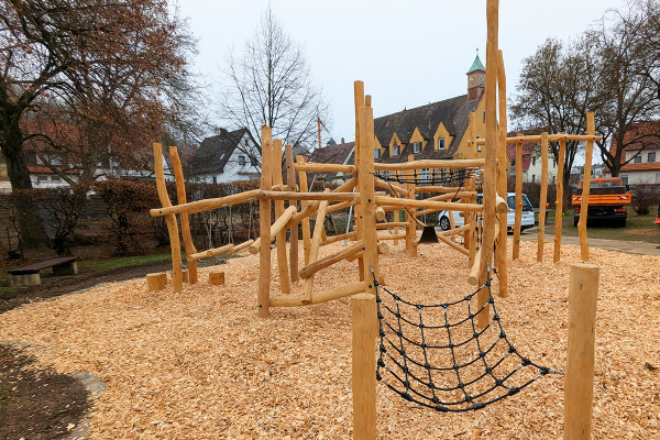Balancier- und Kletterparcours aus Naturholz mit Netz- und Seilelementen auf einem Spielplatz in Nürtingen, mit Kirche und Wohnhäusern im Hintergrund