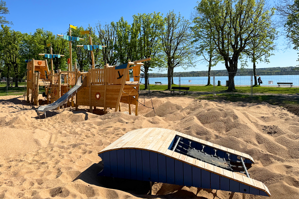 Spielplatz mit großem Holzspielschiff und vorgelagerter Spielwelle im Sandbereich an der Seepromenade, umgeben von Bäumen und mit Blick auf den See.