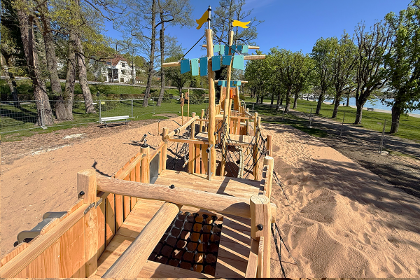 Blick über das Deck eines hölzernen Spielschiffs mit Kletternetzen, Balancierbalken und Masten auf einem Sandspielplatz an der Seepromenade.