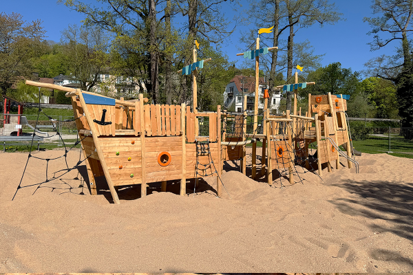 Seitliche Ansicht eines großen Holzspielschiffs mit Kletternetzen, Balancier- und Spielelementen auf einer weitläufigen Sandfläche, umgeben von Bäumen.