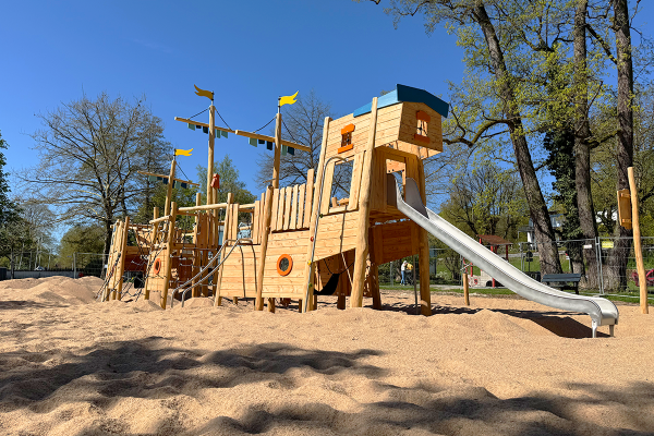 Großes Spielschiff aus Holz mit hoher Rutsche, Kletterelementen und Plattformen auf einem Sandspielplatz unter Bäumen.