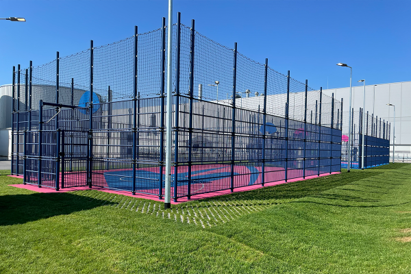 Eingehauste Multisportanlage mit blau-pinkem EPDM-Sportboden und hoher Ballfangzaunanlage auf dem Gelände der Tesla Gigafactory in Grünheide.