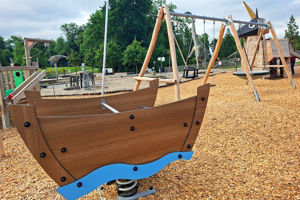 Federwipper in Bootsform und Nestschaukel auf dem Spielplatz im Himmelsgarten Schwäbisch Gmünd. Im Hintergrund ist das Einhorn-Spielgerät aus Naturholz zu sehen, eingebettet in eine großzügige Fallschutzfläche.
