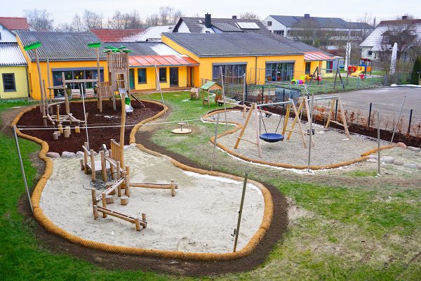 Gesamtansicht des naturnah gestalteten Außengeländes der Kita Karlskron mit Holz-Spielanlage, Wasserspielbereich, Schaukeln und vielfältigen Bewegungsangeboten.