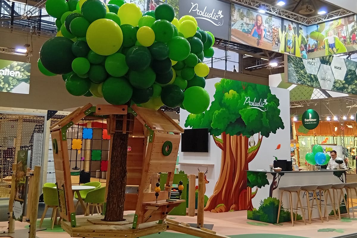 Proludic Messestand mit der Arborea Play® Spielanlage rund um einen Baum und naturinspiriertem Standdesign.